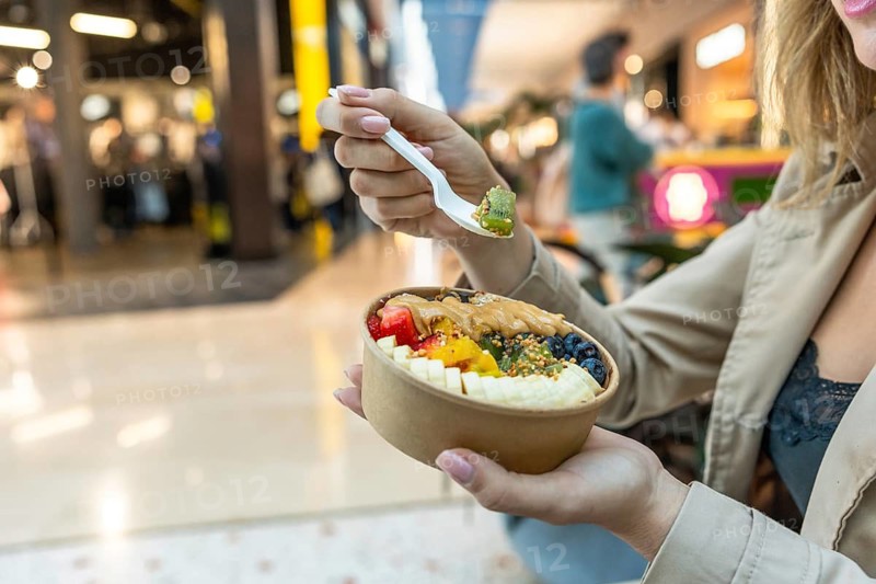 Woman holding biodegradable bowl of colorful acai with kiwi, banana, berries, mango, peanut butter drizzle and bee pollen, enjoying a healthy snack in a modern urban mall setting