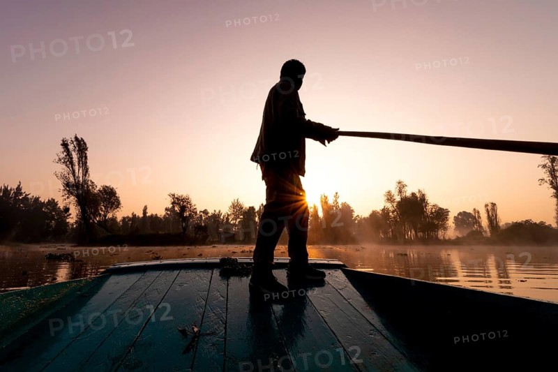 A trajinera boatman is silhouetted against the golden sunrise as he navigates the tranquil canals of Xochimilco, Mexico, using a long wooden pole. A scene of cultural tradition and serenity