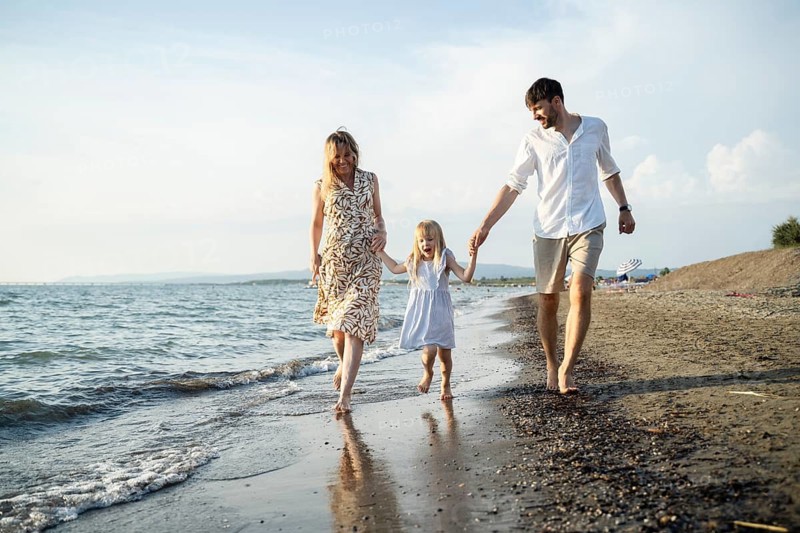 A cheerful family of three enjoys a relaxing walk along the shoreline, holding hands and smiling. The warm evening sunlight casts a golden glow on the peaceful beach setting