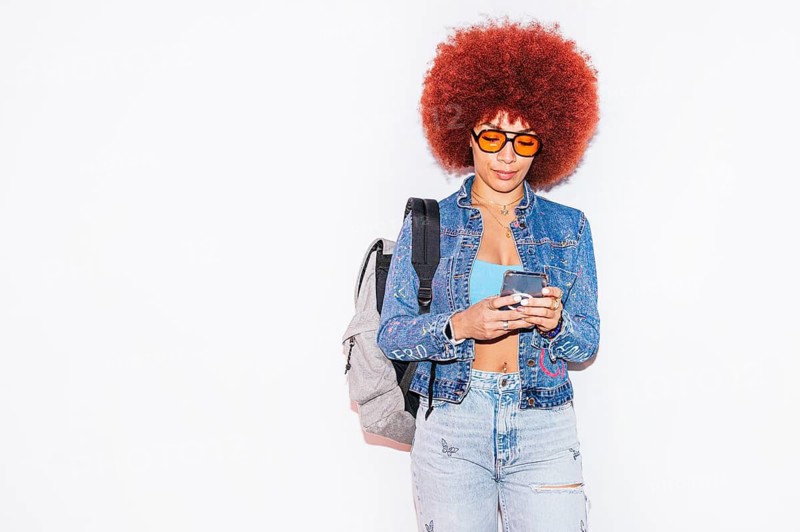 African descendent woman with vibrant afro hair using her smartphone. She sports a denim jacket and jeans, accessorized with backpack and trendy orange sunglasses against a white background