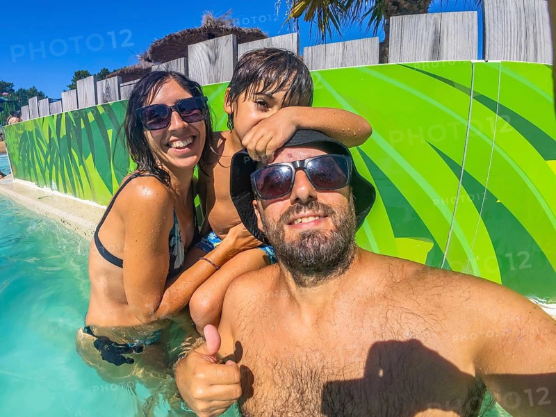 Happy family of threebearded dad in sunglasses and hat, mom in a bikini and their childtaking a cheerful pool selfie while cooling off at a sunny water park resort