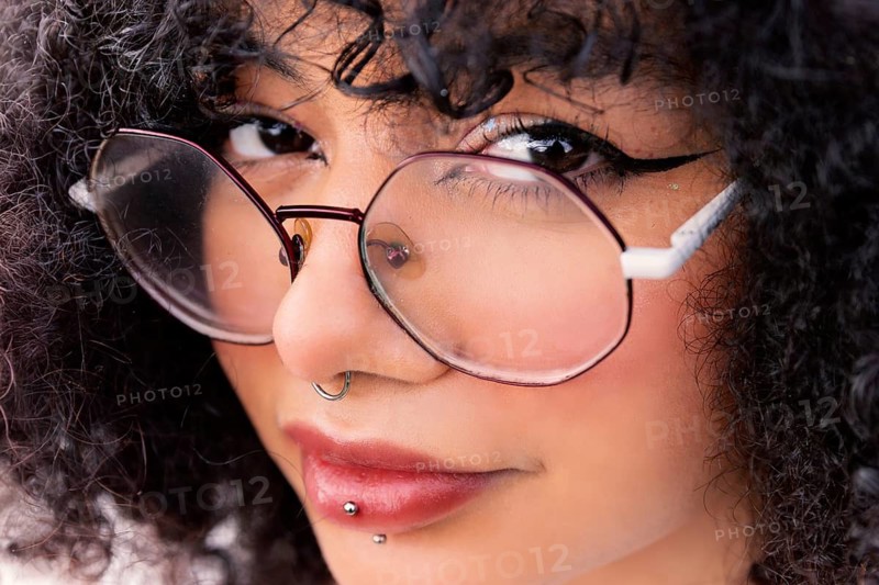 Close-up of a stylish latin woman with large glasses, curly hair, and facial piercings, showcasing an expressive and confident look. Captures modern beauty and individuality