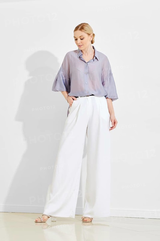 A woman stands confidently in a minimalist space, showcasing a light-colored, oversized shirt and flowing white trousers