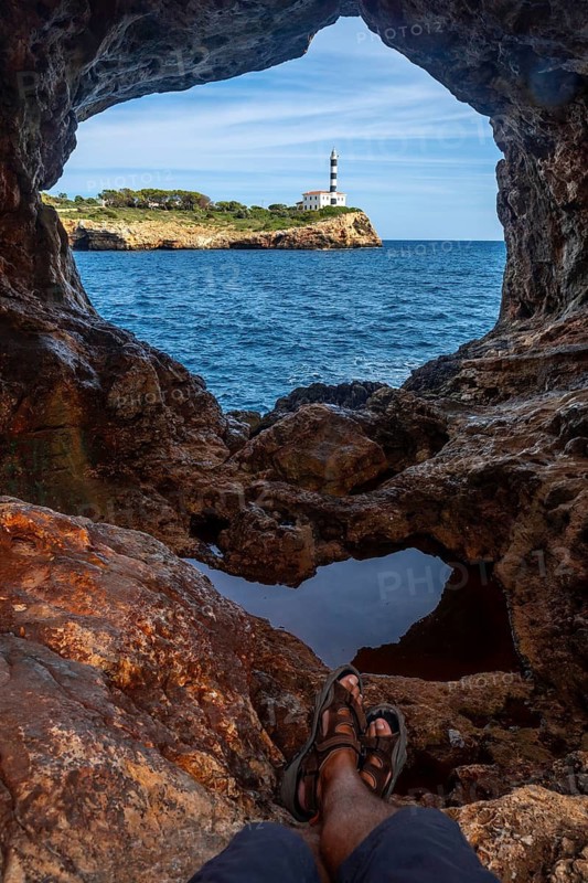Tourist relaxing inside arc natural cave enjoying the view of portocolom lighthouse on a sunny day in mallorca, spain