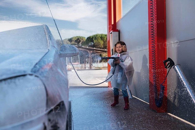 A smiling child in earmuffs and a raincoat playfully washes a car at a self service station, wielding a hose and creating foam in a fun outdoor setting