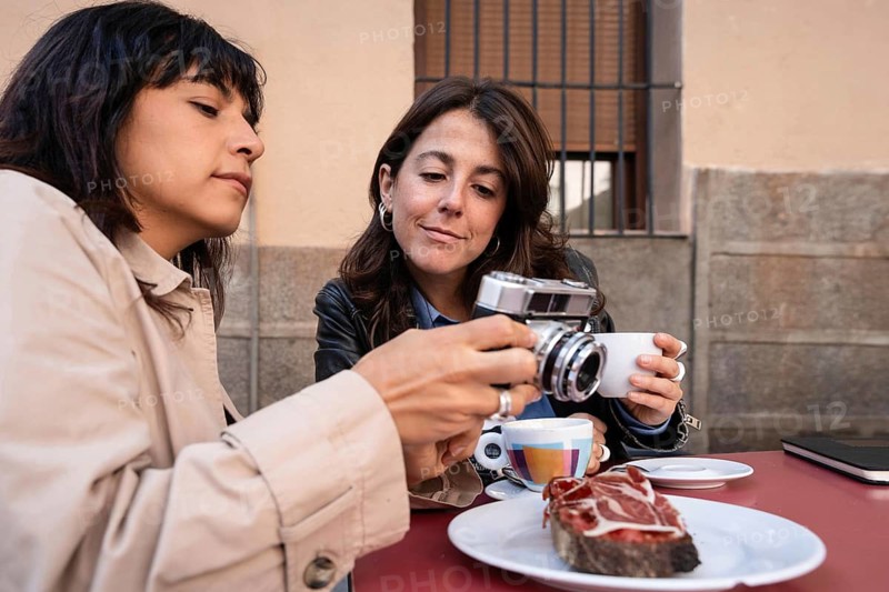 A lesbian couple shares a quiet moment at an outdoor cafe, drinking coffee and looking at a camera. On the table is a traditional Spanish breakfast with ham, which contributes to the cozy atmosphere