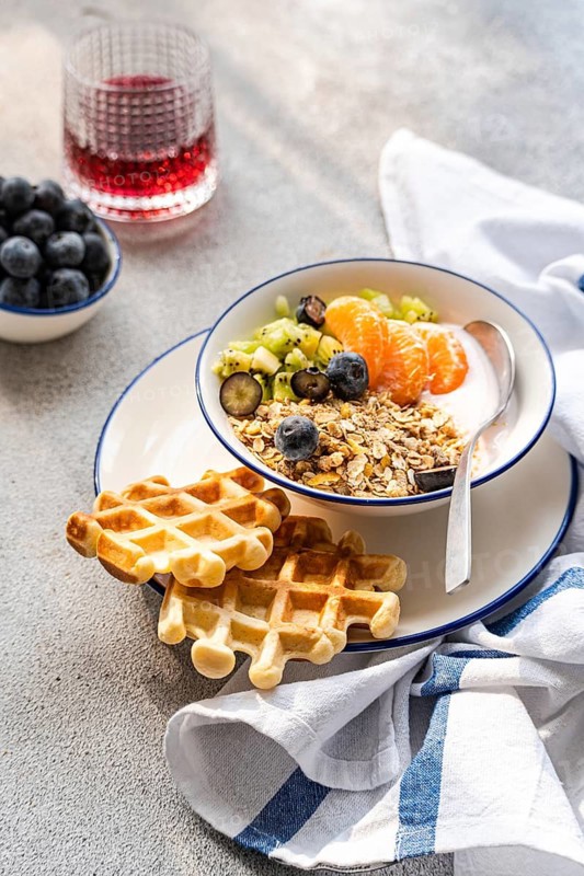 A nutritious breakfast bowl with yogurt, granola, fresh kiwi, blueberries, and tangerine slices. Paired with golden waffles and a vibrant juice, perfect for a morning meal