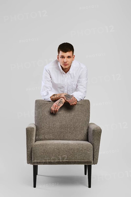 Full length studio portrait of young barefoot man in white shirt and ivory trousers leaning to armchair