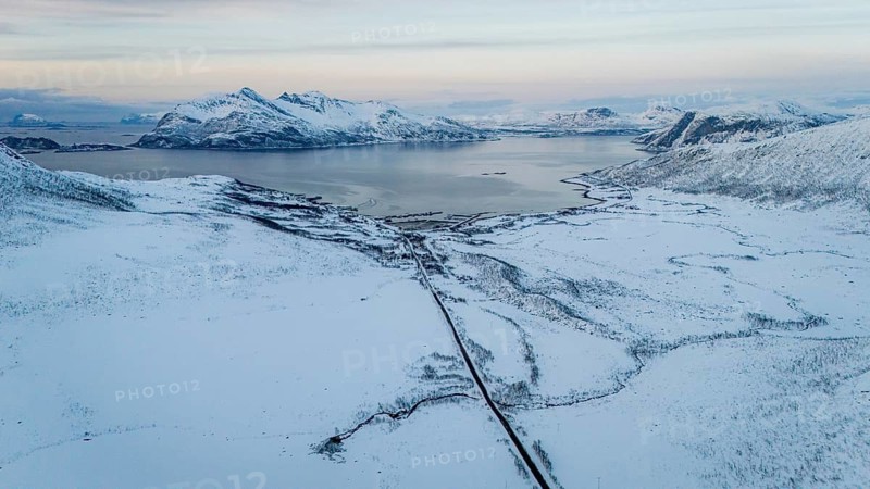 Breathtaking aerial image of a snowy coastal region in Norway, with vast white plains, mountain ranges, and a winding road leading toward calm northern waters at dusk, Kvaløya, Troms, Norway