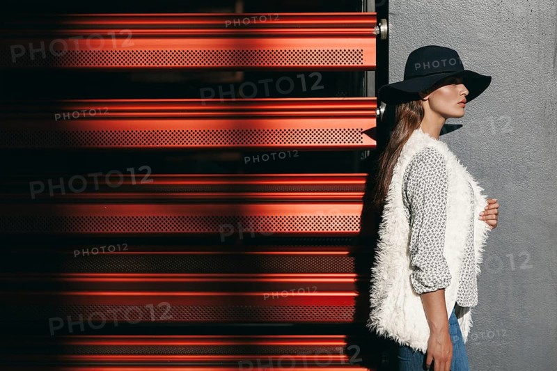 A stylish woman wearing a wide-brimmed hat and textured vest stands against a modern red and gray wall. The striking light and shadow enhance her sophisticated look