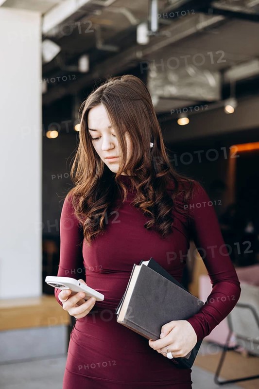 Female student in a maroon dress multitasking, holding books and using a smartphone, ready to study or communicate in an academic setting