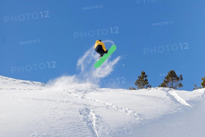 A snowboarder in a yellow jacket and green snowboard catches air on a snowy slope against a backdrop of clear blue sky, showcasing thrilling winter sports action