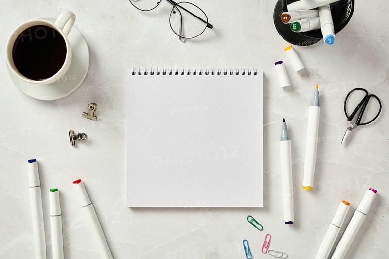 Top view of a white sketchbook surrounded by colorful markers, a cup of coffee, glasses, and office supplies, neatly arranged on a marble table