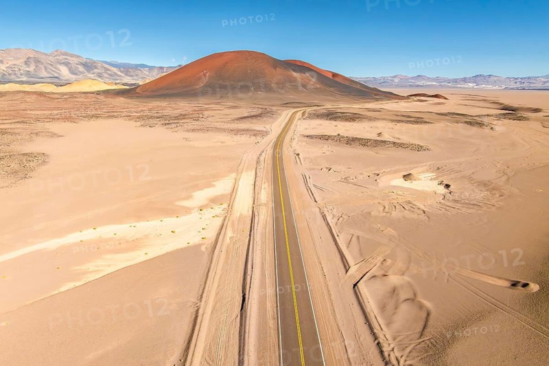 A long, straight road cuts through a vast desert, leading towards a distant, brown hued mountain under a clear blue sky in the argentine Puna, creating a sense of solitude and adventure