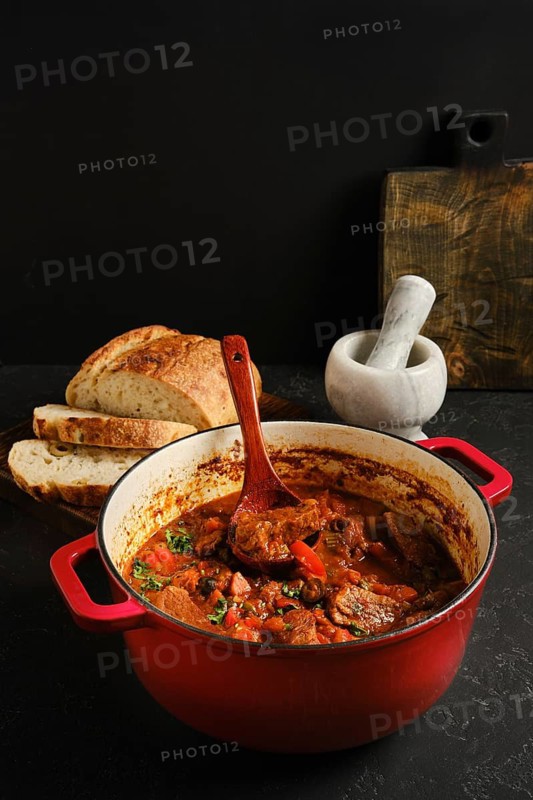 A red cast-iron pot with a hot beef stew . There is a wooden spoon in the pot. Slices of bread sit next to the pot. A mortar is in the background. It is a cozy kitchen scene