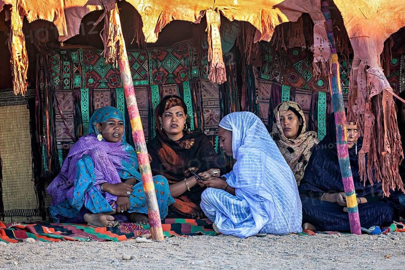 Women sit in a tent and talk to each other, surrounded by colourful fabrics, Veiled woman at the Tamanrasett Festival in the Sahara in Algeria