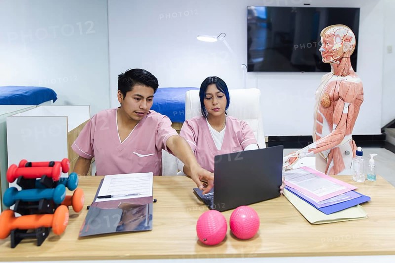 Two physiotherapy professionals in Ecuador discuss treatment plans at a desk. Around them, anatomical models and equipment illustrate a modern clinical setting