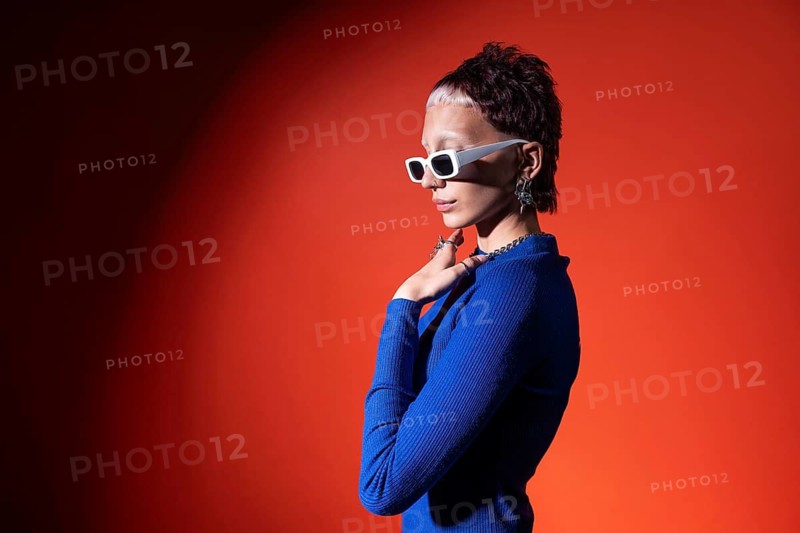 A fashionable young woman in a vibrant blue top poses confidently against a bold red background. She sports stylish white sunglasses and unique accessories, showcasing modern fashion trends