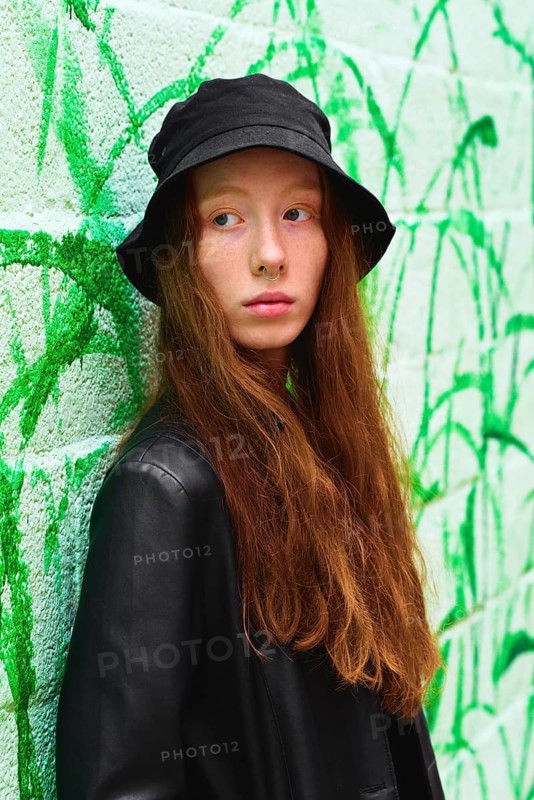 A young woman wearing a black bucket hat and leather jacket poses against a green graffiti-covered wall Her long red hair and calm expression capture a modern street style