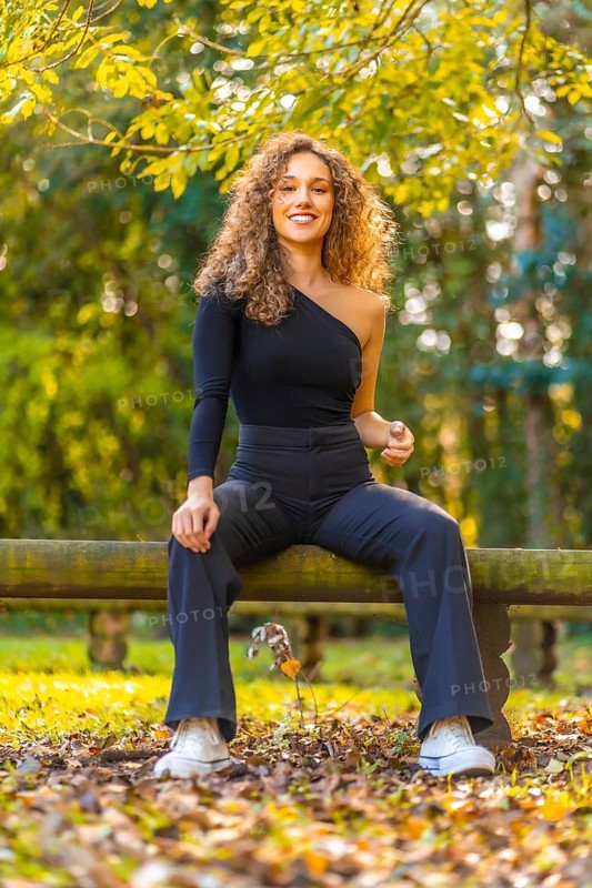 Vertical photo of a young happy woman sitting on a trunk in a park in autumn