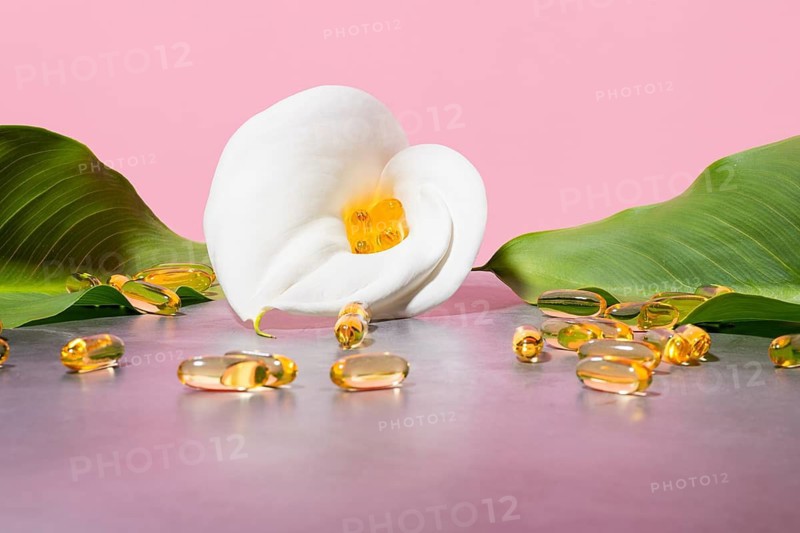 Serene display featuring golden omega-3 oil supplement capsules nestled in a white calla lily, surrounded by lush green leaves against a soft pink background, emphasizing beauty and wellness