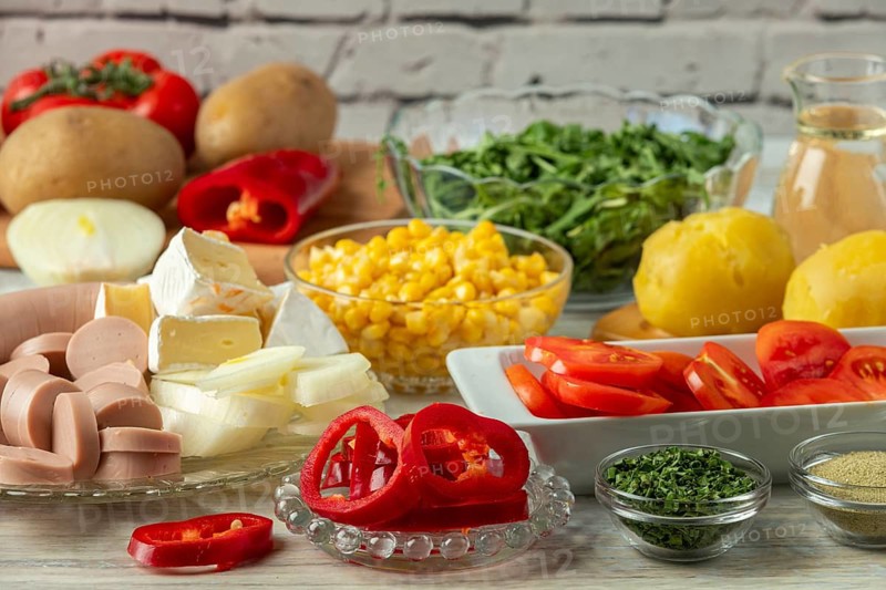 Preparation for salad with potatoes, tomatoes, peppers, sausage, corn, onions, cheese and arugula in a bright environment