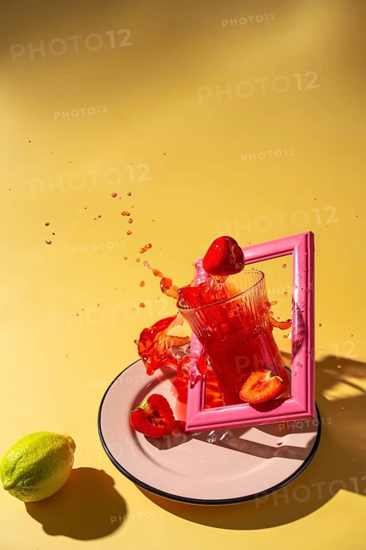 A vibrant image featuring a refreshing strawberry drink splashing out of a glass. The setup includes fresh strawberries and a lemon placed against a yellow backdrop, capturing a lively summer vibe