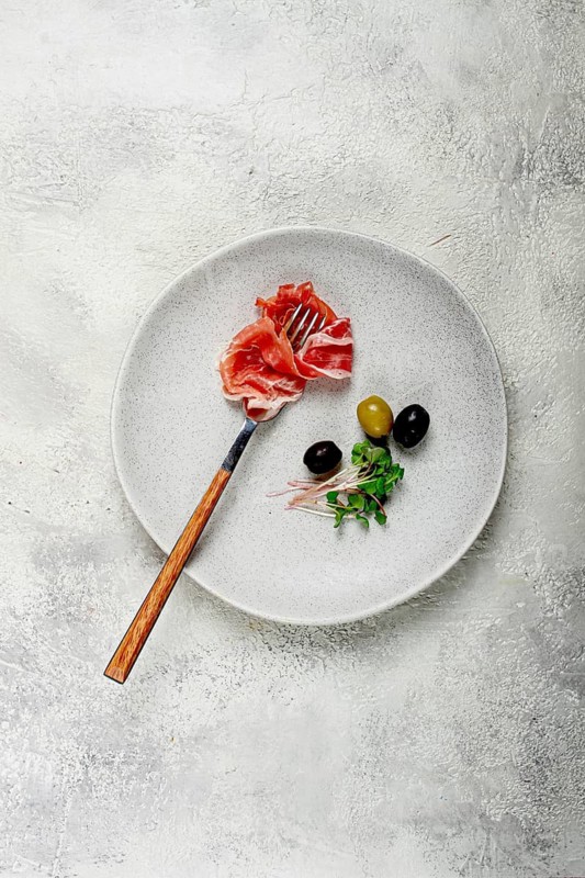 Prosciutto on a fork, top view, olives and micro-greens on a plate, light background, no people