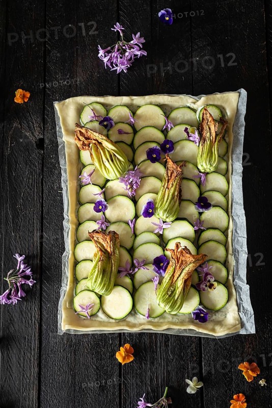A beautifully arranged zucchini and edible flower tart sits on a dark wooden table. The vibrant colors of the flowers contrast with the green zucchini, creating a visually appealing dish