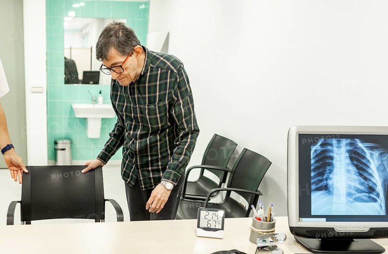 An elderly man in a mental health residence prepares to sit during a medical consultation. The setting includes a doctor's office with an X-ray on the screen, reflecting healthcare and well-being