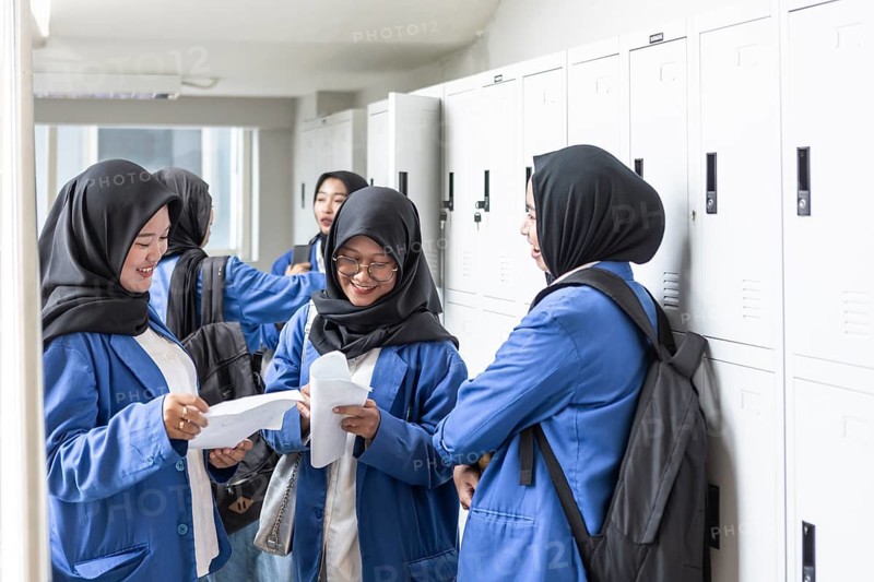 Asian Muslim students in blue uniforms happily share papers in a hallway, standing near lockers. Backpacks and bright smiles suggest a positive return to school atmosphere