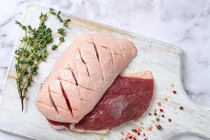 Fresh raw duck breasts, spread on a wooden cutting board, with herbs and spices, on a white marble surface