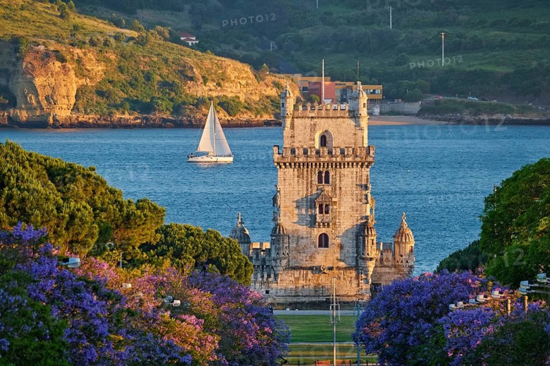 Belem Tower stands majestic against the Tagus River, surrounded by blooming jacaranda trees and sailing boats at sunset