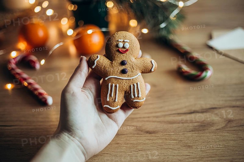 A hand holds a gingerbread man cookie decorated with icing In the background, blurred festive elements like candy canes and oranges create a cozy holiday atmosphere