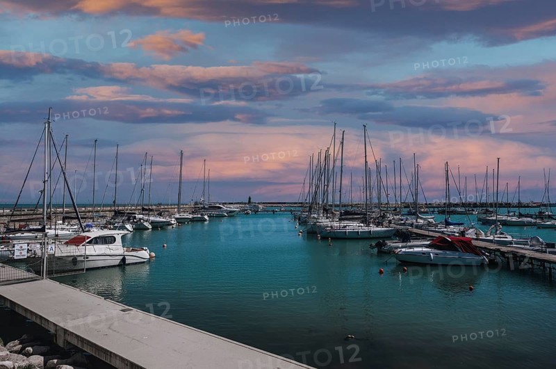 A stunning marina under a vibrant sunset sky in Civitanova Marche, Italy. The scene captures the serene waters and moored sailboats, reflecting pastel hues from the clouds