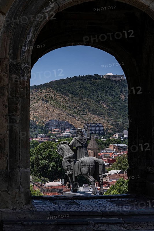 An equestrian statue framed by an archway overlooks Tbilisi's blend of historical and modern architecture, backed by lush green hills under a clear blue sky