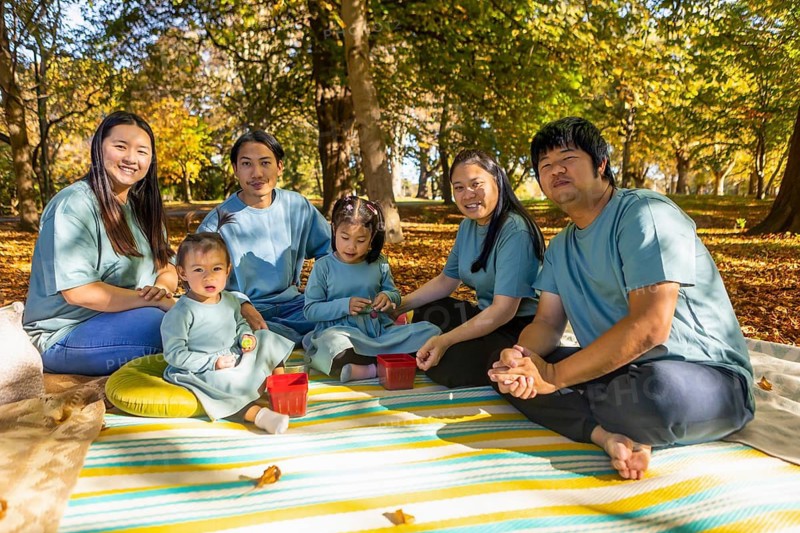 An Asian family enjoys a picnic during autumn, surrounded by vibrant foliage. They sit on a colorful blanket, smiling and bonding in the serene park setting