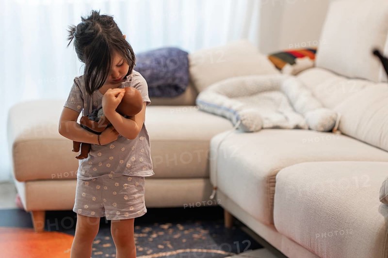 A young girl lovingly embraces her doll in a cozy living room, capturing a tender moment. The scene reflects a warm family atmosphere, expressing love and comfort
