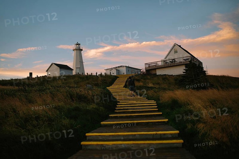 Back view of an unrecognizable man ascending a yellow painted staircase towards the historic Cape Spear Lighthouse in Newfoundland, under a dusky sky