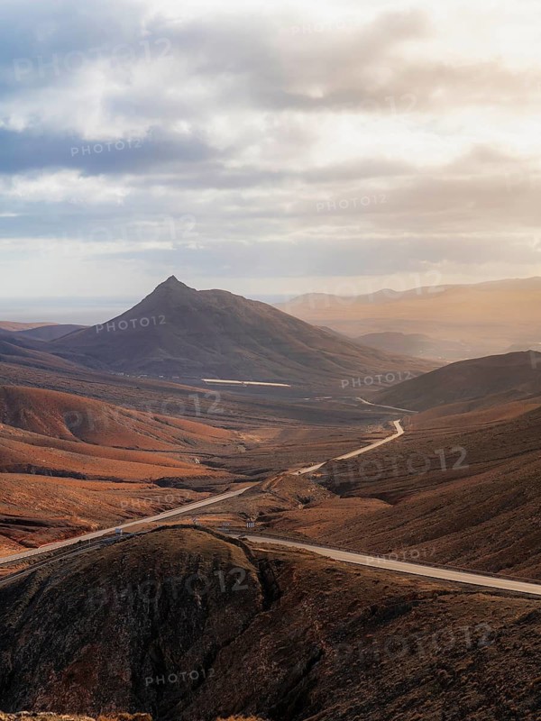 A breathtaking view of a winding road cutting through the rugged mountains of Fuerteventura. The warm sunlight and dramatic clouds highlight the island's vast, dry, and volcanic terrain