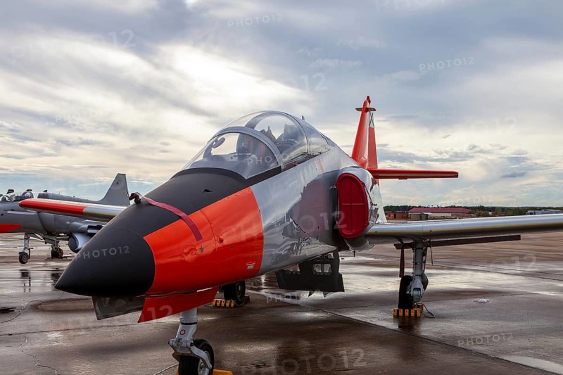 A sleek military training jet with red and black markings is stationed on the wet runway at an air base. The aircraft's aerodynamic design highlights its speed and agility for pilot training