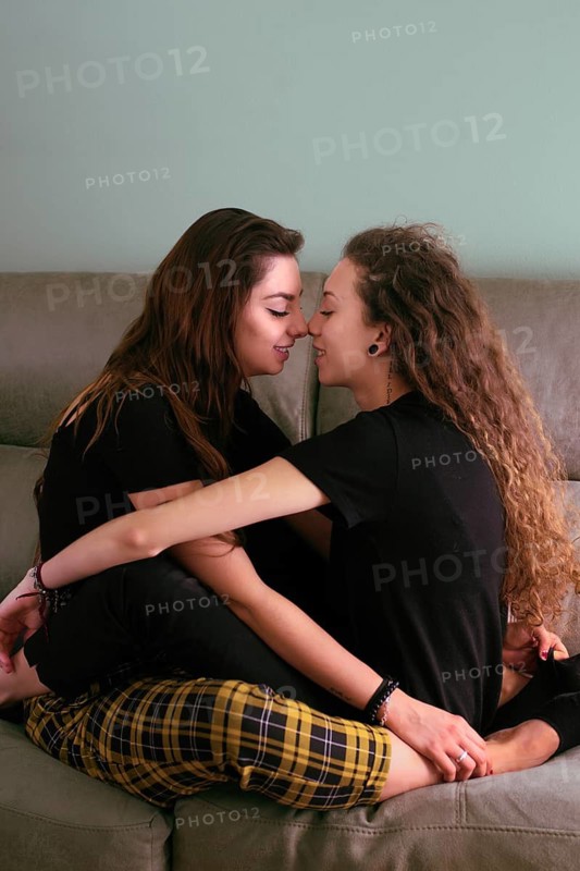 A tender moment as a female gay couple embraces on a sofa, touching noses and smiling gently at each other in a loving gesture, conveying affection and closeness in their private space