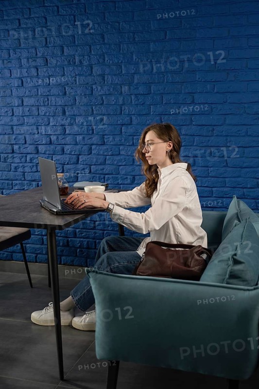 A woman in casual attire works on her laptop in a modern cafe with a vibrant blue brick wall. The scene suggests productivity and a relaxed work environment