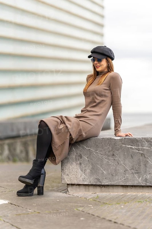 Stylish woman posing wearing a beige turtleneck dress, newsboy cap, sunglasses and high heel boots