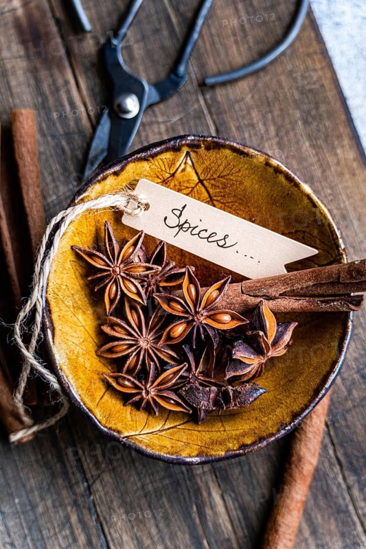 Top view of star anise and cinnamon sticks in a decorative yellow bowl, with a Spices written tag, surrounded by rustic tools on a wood surface