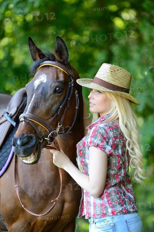 Woman (cowgirl) in a straw hat, plaid shirt and denim shorts stands beside a polish arabian horse on a field, Bavaria