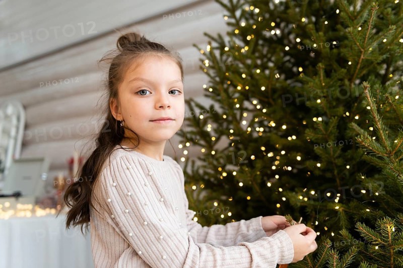 A young girl with a gentle expression is seen decorating a lush Christmas tree, which is brightly lit with tiny golden lights. She wears a cozy, textured sweater and looks directly at the camera, her blue eyes clear and focused