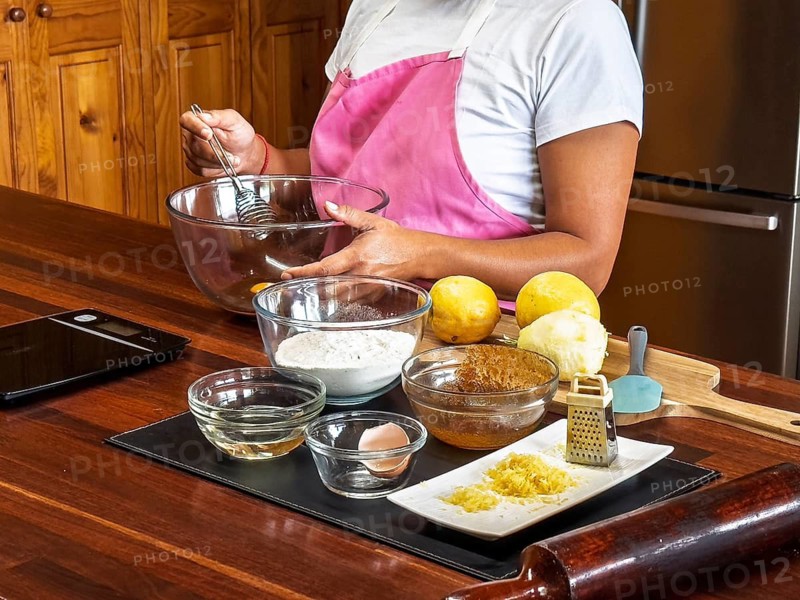 A woman wearing a pink apron prepares ingredients for baking lemon cookies in a kitchen, surrounded by various baking tools and ingredients such as flour, lemons, and a grater on a wooden counter