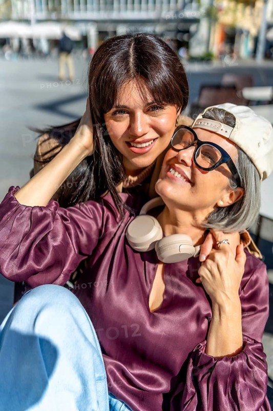 Two smiling women, a mother and her adult daughter, embracing and enjoying a moment of family bonding and connection together in an urban outdoor setting