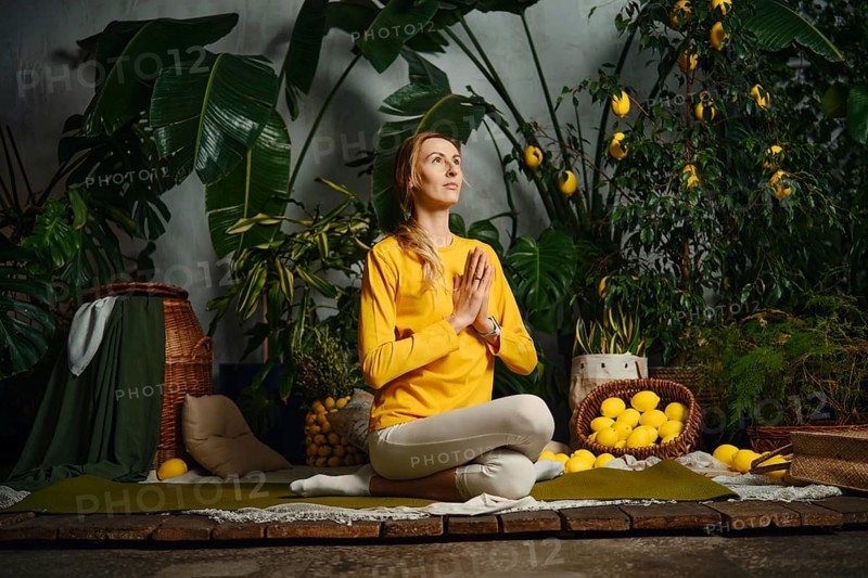 A woman sits on a mat in a garden filled with plants and lemons. She holds her hands together in a pose. The sunlight shines in, creating a bright and inviting scene for yoga practice